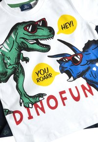 Bílý tričko s motivem zeleného Tyrannosaura a modrého Triceratopse se slunečními brýlemi, textovými bublinami a nápisem "DINOFUN" tučným červeným písmem.
