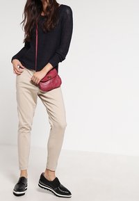 Marinblå stickad tröja, beige avsmalnande byxor och svarta snörskor. En liten röd väska över axeln ger en färgklick.