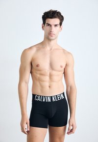 Crne pamučne bokserice s bijelim logom "CALVIN KLEIN" na pojasu, prilagođeni dizajn, glatka tekstura, nosi ih muški model.