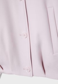 Světlounce růžová látka se dvěma kulatými knoflíky a vertikálním šlicem kapsy na pravé straně oděvu.