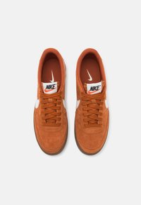 Narancssárga velúr Nike sneaker, fehér swoosh-zal. Barna gumitalppal és narancssárga hálós béléssel, fűzős kialakítással és párnázott gallérral.