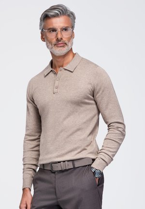 Un homme aux cheveux argentés, barbe et lunettes, pose en pull polo beige, pantalon gris, ceinture et montre-bracelet sur un fond uni.