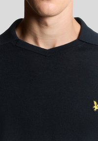 Pull collo V lavorato a maglia di colore blu navy, con un piccolo logo giallo ricamato sul lato sinistro del petto. Texture liscia.