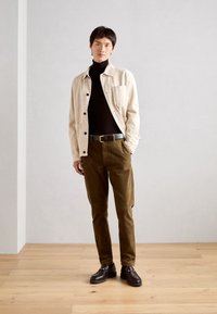 Beige canvasjacka över en svart polotröja, i kombination med olivgröna kordbyxor och mörka loafers. Minimalistisk design, funktionella fickor.