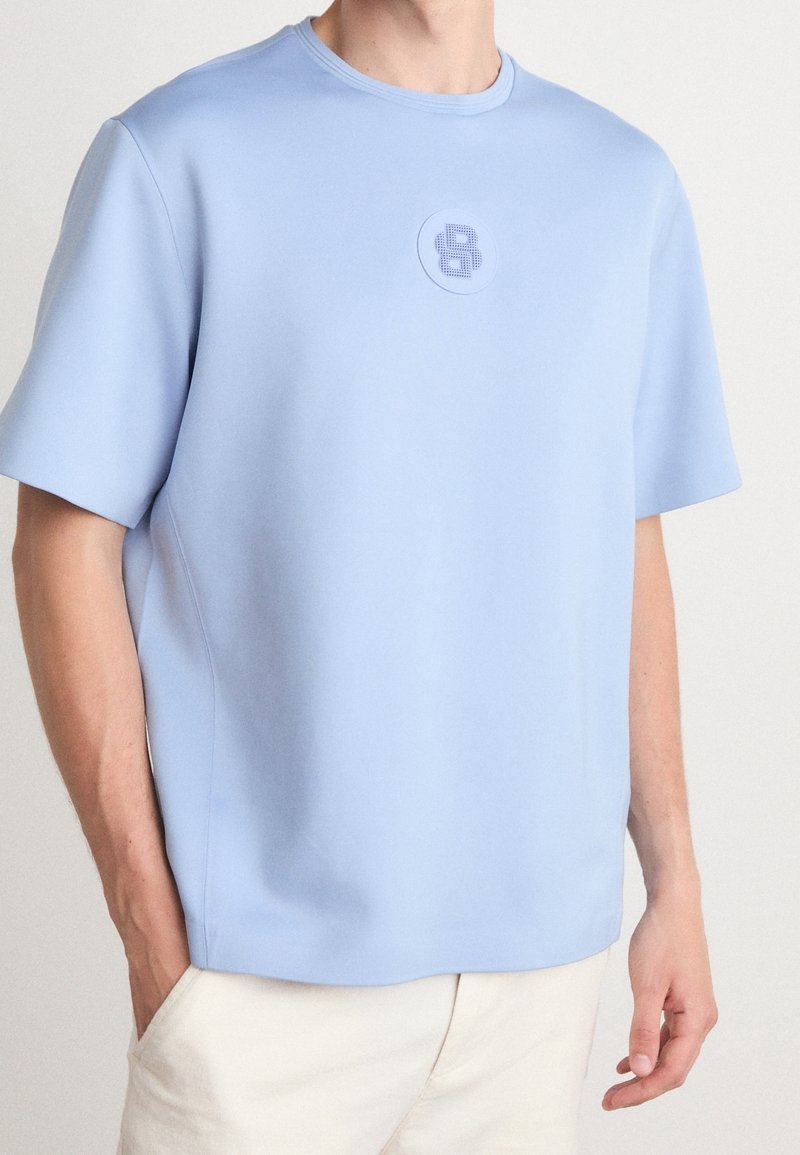 T-shirt bleu clair à manches courtes en tissu lisse, avec un col rond et un logo circulaire sur la poitrine.