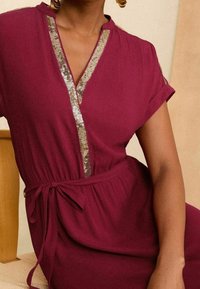 Viininpunainen mekko lyhyillä hihoilla, V-pääntiellä ja kullanvärisellä paljettiompeleella edessä. Muotoiltu vyötärö vyöllä, pehmeä tekstuuri.