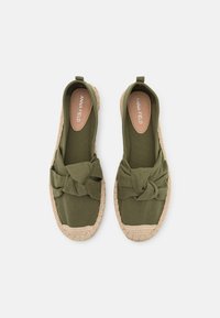 Espadrilles en tissu vert olive avec un détail noué à l'avant et une semelle en jute tressé. Texture lisse, design enfiler, et talon renforcé.