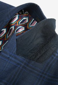 Blazer de cuadros azul marino con un cuello de fieltro negro en contraste y un forro de patrón geométrico colorido. Detalle de costura visible.