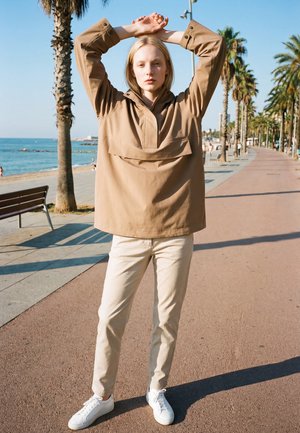 Ung person i beige jakke og bukser, der strækker armene over hovedet på en solrig palmebevoksede havnepromenade.