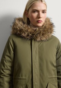 Parka verde oliva con una gran capucha forrada de piel marrón y un collar acanalado beige. Cuenta con dos bolsillos frontales y cierre de cremallera.