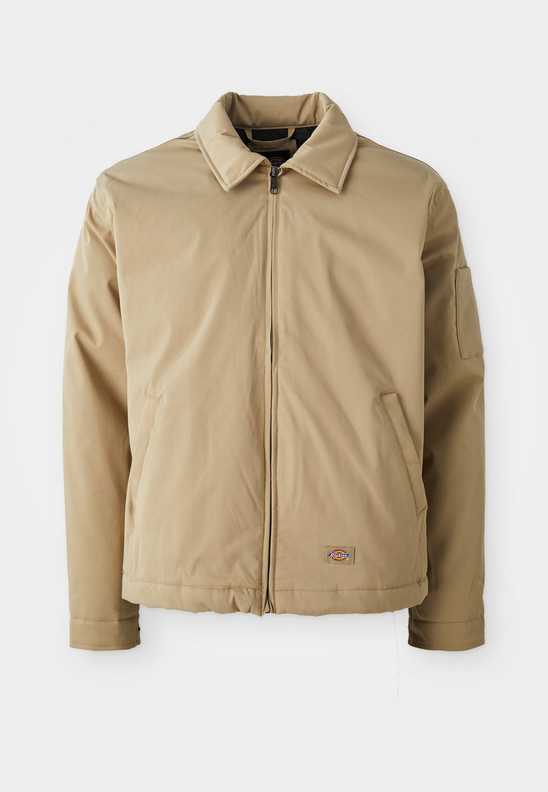 Veste beige zippée avec un col pointu, deux poches avant et un petit logo près de l'ourlet, présentée sur un fond blanc.