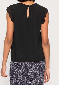 Schwarze kurzärmelige Bluse mit gerafften Kanten, die über einen Schlüssellochverschluss im Rücken verfügt. Der Stoff ist leicht und hat ein durchsichtiges florales Muster am Saum.