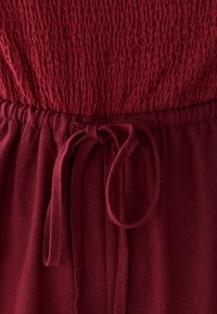 Obleka bordo barve z zgornjim delom z naborki in pasom z vrvico. Tkanina ima teksturiran videz in rahlo raztegljivost.