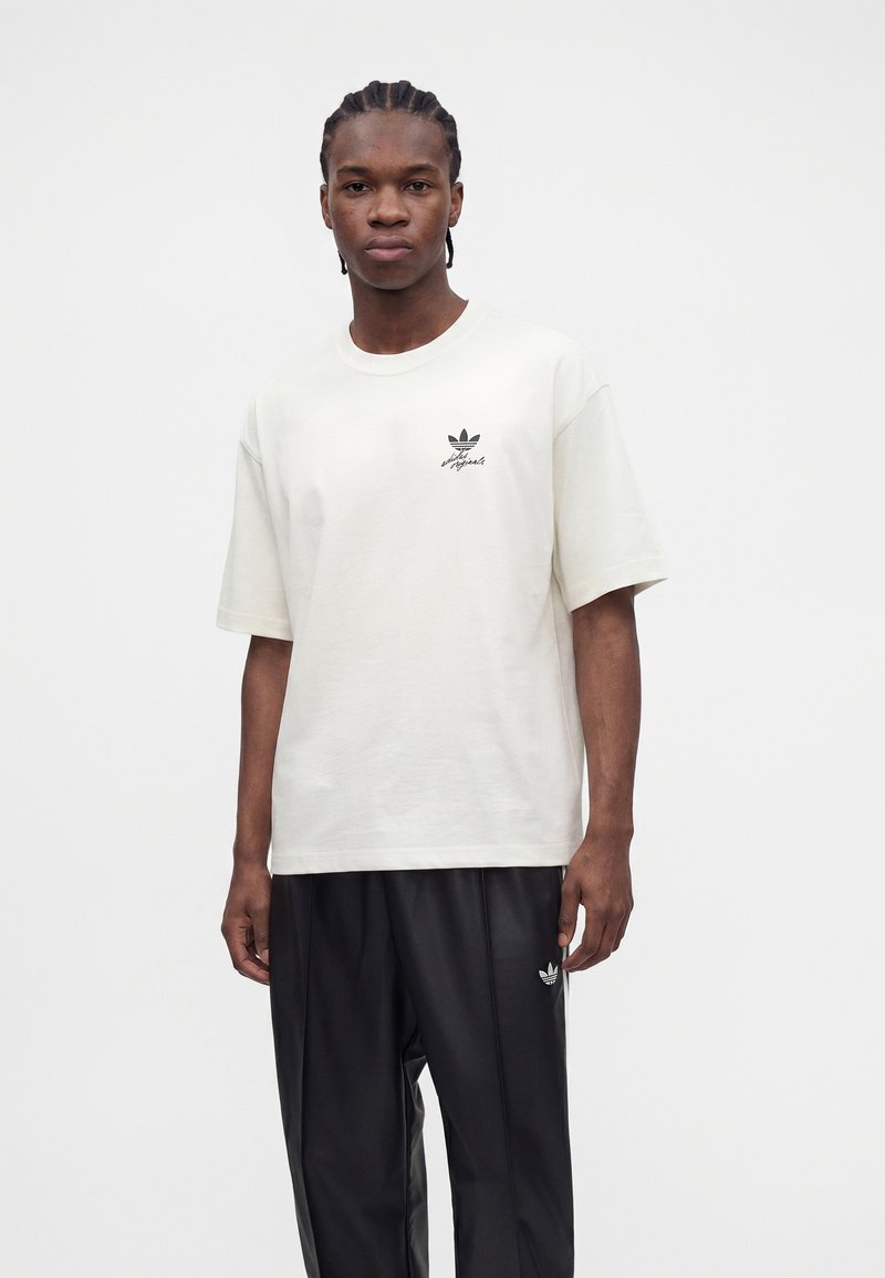 Jeune homme aux cheveux tressés, portant un t-shirt blanc Adidas et un pantalon noir Adidas, debout devant un fond clair uni.