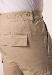 Beige shorts lavet af struktureret stof med elastisk talje og baglomme. Har broderet logo på bagsiden.