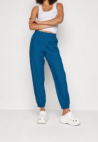 Modré, lehké joggingové kalhoty s elastickým pasem, nařasenými manžetami a bočními kapsami, ve spojení s bílým tílkem a bílými dřeváky.