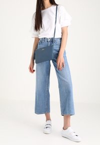 Piccolo borsa a tracolla blu, in pelle liscia, con zip, tracolla regolabile, con un accento a frange. Abbinata a una maglietta bianca e jeans a vita alta.