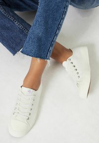 Baskets en toile blanches avec des bouts ronds, œillets en métal, lacets plats et semelles texturées, associées à un jean bleu avec un ourlet effiloché.