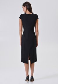 Donna che indossa un vestito nero aderente al ginocchio con maniche corte, zip sul retro e spacco posteriore, abbinato a scarpe nere con tacco alto.