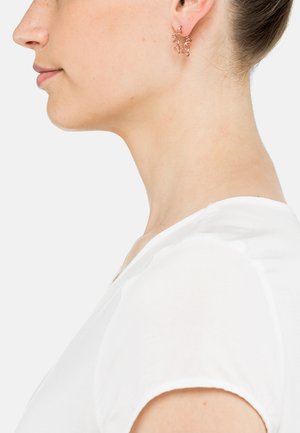 Roségoldene Ohrringe mit einem zarten, verflochtenen Blättermuster, von der Seite betrachtet, getragen von einer Person in einem weißen Hemd.