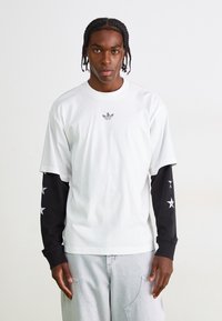 Tricou alb cu un mic logo negru pe piept, asortat cu mâneci lungi negre împodobite cu modele albe de stele.