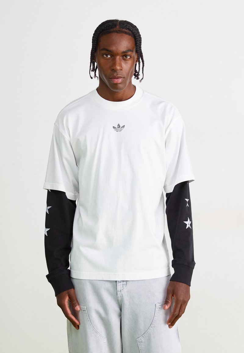Tricou alb cu un mic logo negru pe piept, asortat cu mâneci lungi negre împodobite cu modele albe de stele.