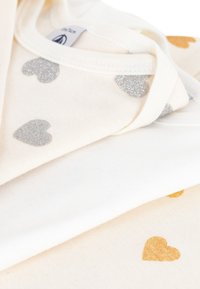Vita och krämfärgade tygskikt med metalliska guld- och silverhjärtan. Mjuk textur, utformad för komfort och lager-på-lager.