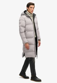 Abrigo largo acolchado de color gris claro con capucha, forro negro, cremalleras laterales y cremallera frontal. Lleva puesto un jersey de punto texturizado tipo cuello alto y jeans oscuros.