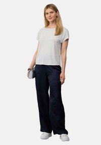 Witte top met korte mouwen, marineblauwe wijde satijnen broek en witte sneakers; model houdt een kleine geprinte clutch met ketting vast.