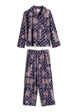 Femme aux cheveux courts bruns portant une chemise boutonnée avec un motif à carreaux bleu et bordeaux et imprimé léopard, regardant sur le côté.