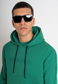 Sudadera verde con un frente texturizado, cordones negros y la palabra "Morato" en relieve. Usada con gafas de sol cuadradas negras.