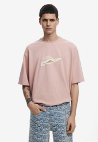 Kortærmet pink T-shirt lavet af bomuld, med et design af revet papir i midten. Model iført blå teksturerede shorts.