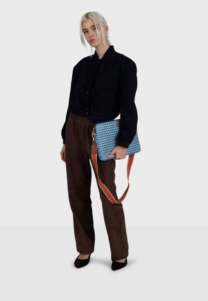 Bolso de hombro azul con estampado y correa marrón, sostenido por una modelo con una chaqueta corta negra y pantalones de ante marrones, de pie frente a un fondo claro.
