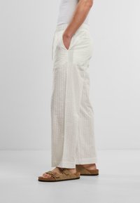 Ľahké, krémové ľanové nohavice s textúrovanými, vertikálnymi pruhmi, elastickým pásom a bočnými vreckami. Skombinované s hnedými šľapkami na slip-on.