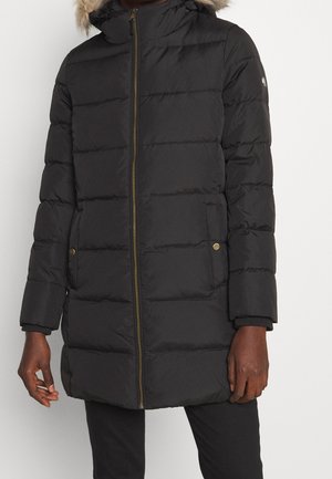 Długa czarna pikowana zimowa kurtka z frontowym zamkiem błyskawicznym, dwoma przednimi kieszeniami na zatrzaski oraz kapturem obszytym futrem, noszona przez osobę.