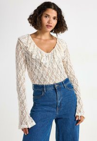 Donna con capelli ricci che indossa una blusa bianca di pizzo a maniche lunghe con rouches e jeans blu a vita alta, in piedi contro uno sfondo neutro.