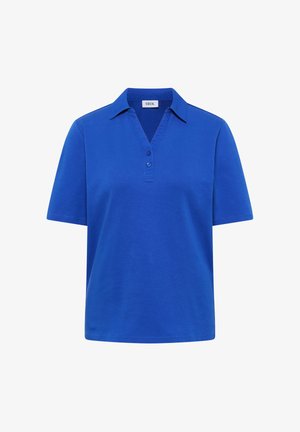 Femme debout contre un mur beige, portant une chemise bleue à col et manches courtes, un jean bleu clair raccourci et des chaussures beiges à enfiler.