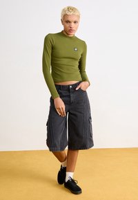 Top aderente a maniche lunghe verde oliva con un piccolo logo, abbinato a pantaloncini neri stile cargo fino al ginocchio e scarpe da ginnastica nere con calze bianche.
