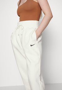 Žena oblečená v bílých Nike teplácích s vysokým pasem a šňůrkou, s rukama v kapsách, a bezrukávovém hnědém topu na jednobarevném pozadí.