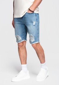Mand iført lyseblå revnede denimshorts, hvide sneakers, hvide sokker og en beige t-shirt, stående foran en ensfarvet baggrund.