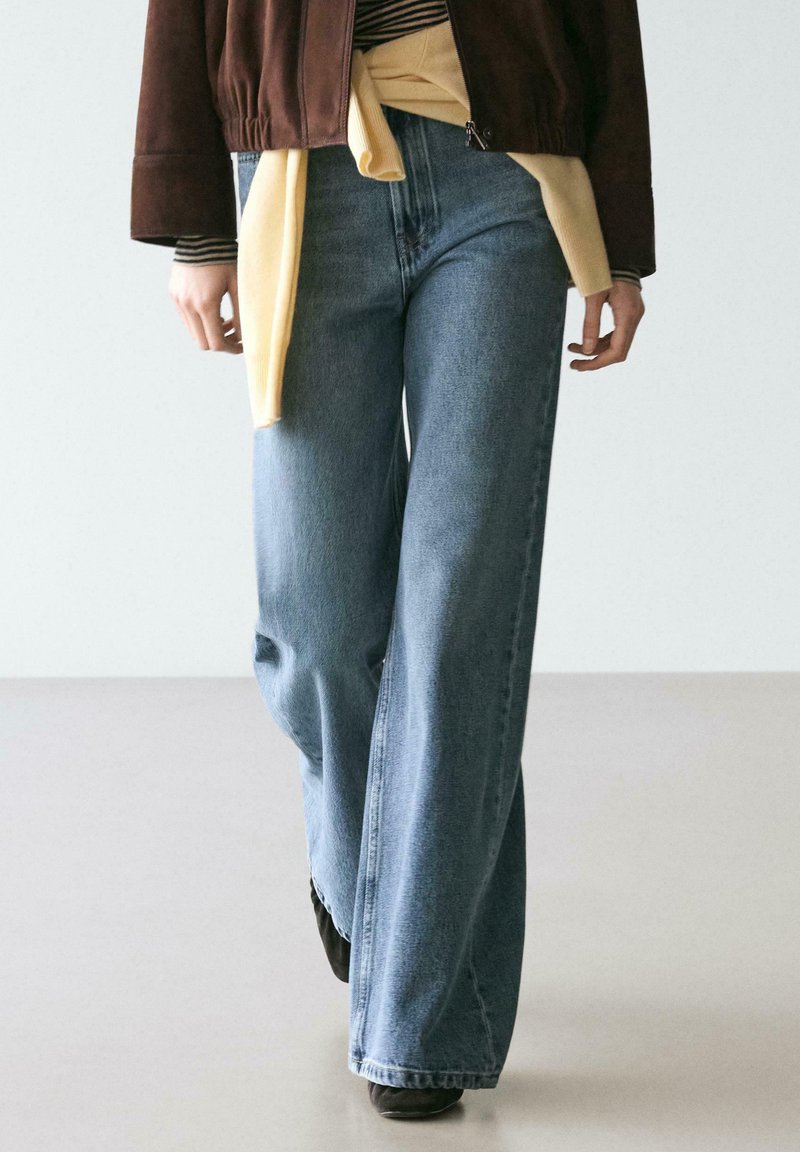 Weit geschnittene Jeans aus hellem Denim mit subtilen Verfärbungen. Kombiniert mit einer braunen Wildlederjacke und einem gelben Pullover, der in der Taille gebunden ist.