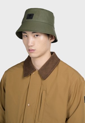Olivgrön bucket hat med en texturerad yta och svart patch. Bärs med en brun kordflöjelkragejacka med tryckknappar och ficka.