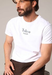 T-shirt en coton blanc avec "Tokyo Deeluxe" imprimé en bleu marine, col rond, manches courtes et coupe décontractée. Pantalon marron foncé visible.