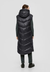 Schwarze Steppweste mit Kapuze, die ein Zickzackmuster, glänzende Textur und lange Länge aufweist, getragen über einem grünen Pullover und dunklen Hosen.