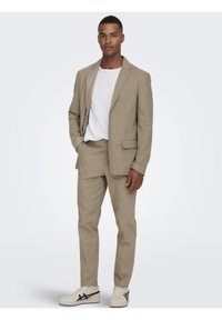 Traje de color beige claro hecho de tejido texturizado, que incluye una chaqueta de un solo botón con solapas de muesca, pantalones a juego y zapatillas blancas.