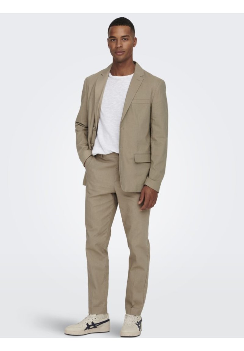 Traje de color beige claro hecho de tejido texturizado, que incluye una chaqueta de un solo botón con solapas de muesca, pantalones a juego y zapatillas blancas.