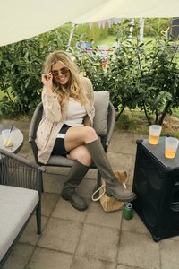 Botas de goma verdes hasta la rodilla, chaqueta beige sobre una blusa blanca, pantalones cortos negros, sentado en una silla de mimbre con un cojín gris, entorno al aire libre.