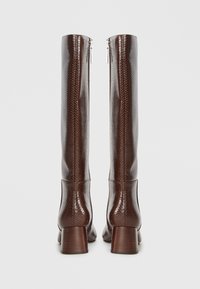 Paire de bottes hautes en cuir marron avec un motif texturé, talons blocs et fermetures éclair à l'arrière sur un fond clair uni.