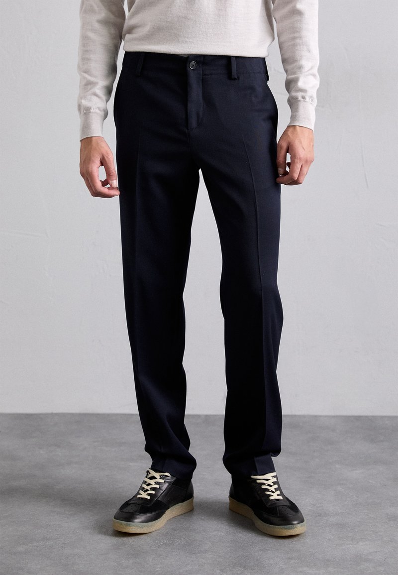 Pantaloni blu navy realizzati in tessuto liscio, con una vestibilità slim, tasche frontali e chiusura classica con bottone. Indossati con sneakers nere.