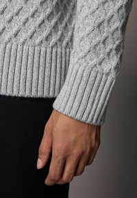 Grauer Strickpullover mit Rautenmuster, geripptem Bündchen und weicher Textur, über dunklem Stoff getragen, der die Handdetail zeigt.